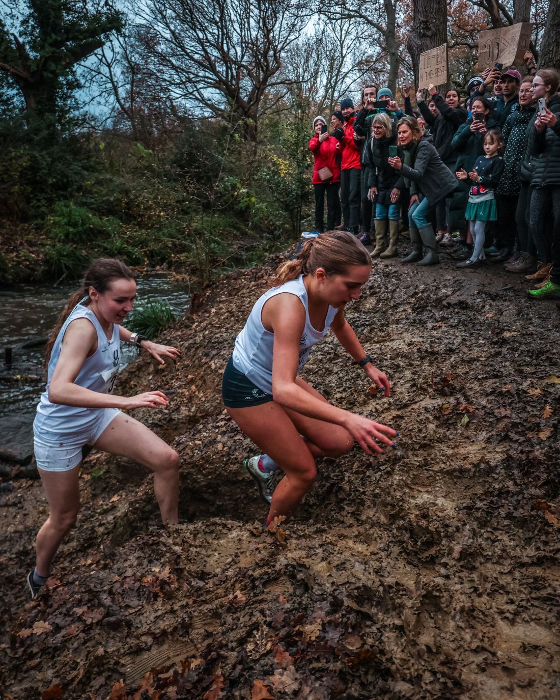 Two Ladies’ runners scrambling up the muddy bank out of the brook as spectators cheer