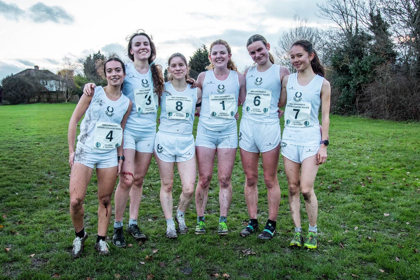 Six Cambridge Ladies’ team runners in race vests stand arm in arm on the grass.